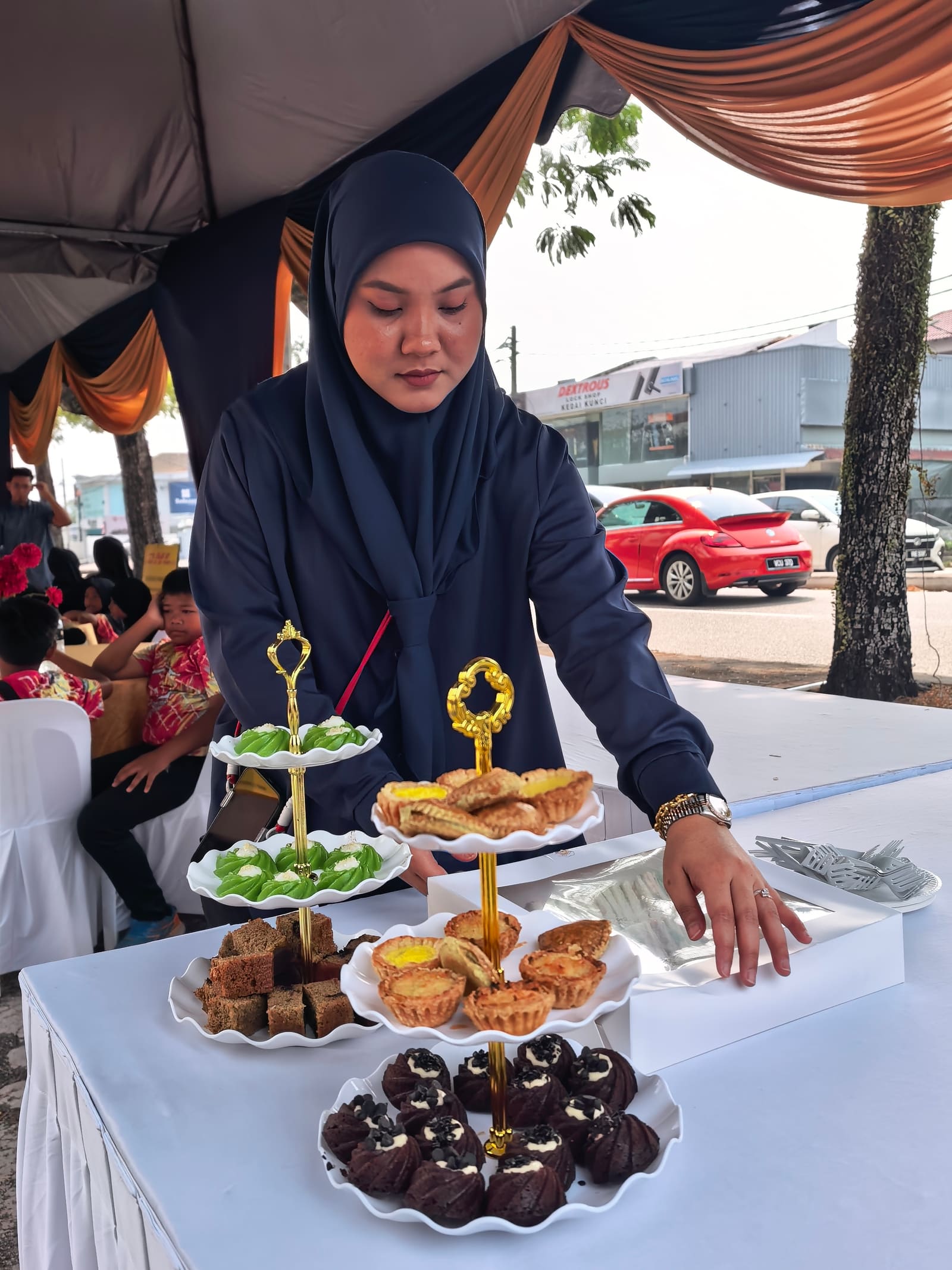 A three-tier stand of kuih, egg tarts and cupcakes being arranged on the street-side tea table, with the marquee going up behind